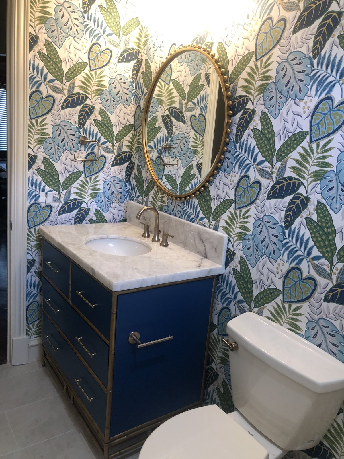 Guest bathroom with botanical wallpaper, a navy vanity and a brass mirror