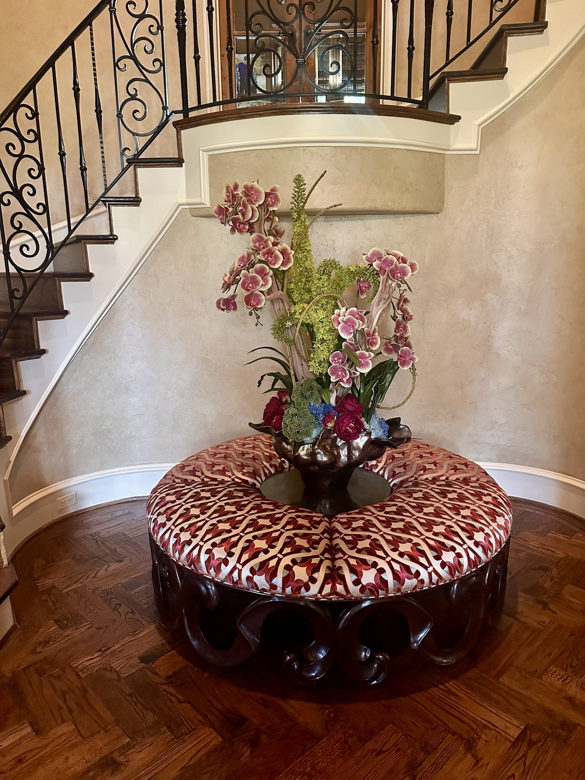 Round tufted ottoman with a tall custom floral arrangement beneath a curved staircase